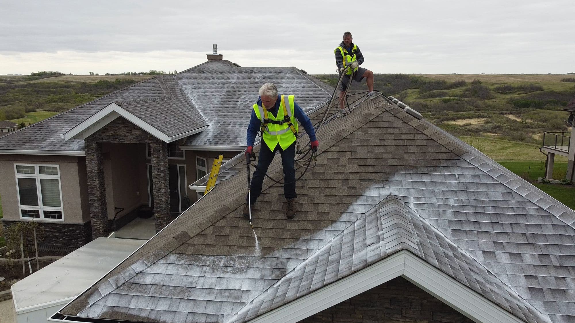 Two roof technicians in high-visibility vests applying rejuvenation treatment to an asphalt-shingle roof