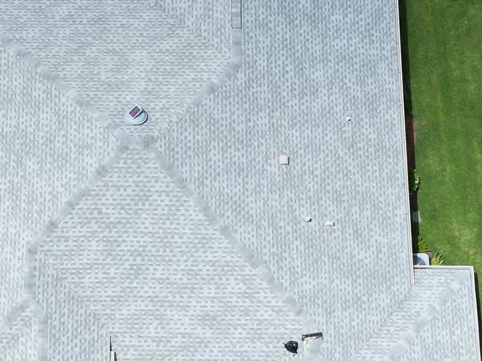 Top-down drone view of a light gray asphalt-shingle roof before treatment, with a faded, chalky surface